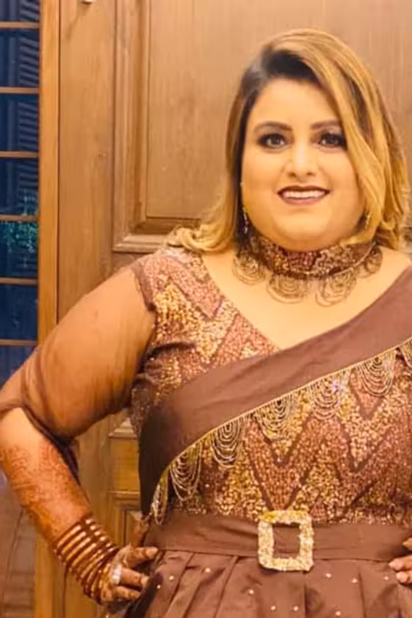 A woman in a long embroidered brown outfit, photographed indoors, hand on her hip